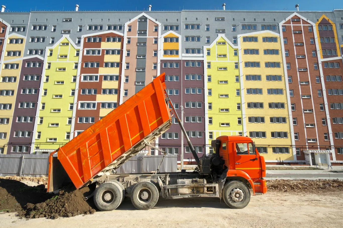 Bright orange dump trailer unloading soil at a construction site near colorful apartment buildings, illustrating a typical urban job site scenario for dump trailer rental in Seattle.