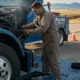 A mechanic in work gloves and a jumpsuit performs reliable semi truck repair on a blue truck by the side of a road, with tools laid out on a blue tarp. In the background, a tow truck from "Williams Diesel" is visible under a clear sky, suggesting a service that also offers reliable semi truck repair in Seattle and surrounding areas.