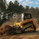 A yellow CAT compact track loader is actively pushing a pile of dark brown mulch on a sunny day, with tall pine trees in the background, making it an ideal visual for a compact track loader rental service.