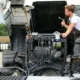 A male mechanic in grey overalls and a white t-shirt is diligently working on the open engine of a large white truck, holding a wrench, symbolizing the detailed work involved in heavy duty truck repair.