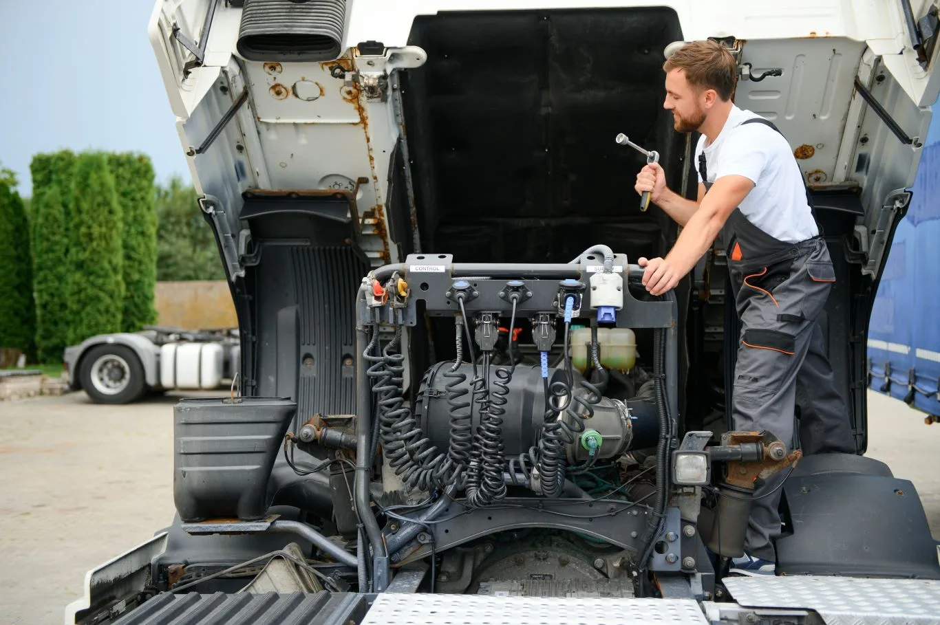 A male mechanic in grey overalls and a white t-shirt is diligently working on the open engine of a large white truck, holding a wrench, symbolizing the detailed work involved in heavy duty truck repair.