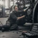 A mechanic, wearing glasses and work clothes, is kneeling on the ground while working on the engine of a large vehicle in a garage, with a toolbox full of wrenches and tools open in the foreground. The image highlights diesel repair services.