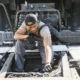 A male mechanic in work overalls and a baseball cap is crouching over the open engine bay of a large truck, inspecting and working on the wiring and components. This image represents diesel truck repair services.