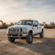 Ford F350 Truck Rental displayed in an open industrial lot, showcasing a sturdy white Ford F350 parked near construction equipment under a soft evening sky.