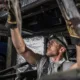 A skilled heavy truck mechanic inspecting components beneath a large vehicle, wearing protective gloves and safety gear while checking the undercarriage.
