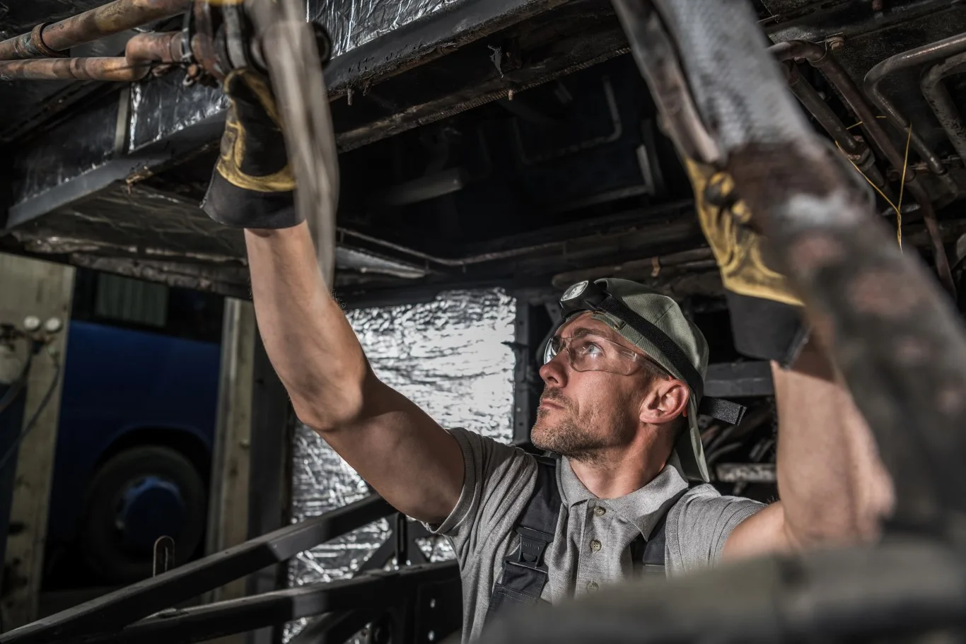 A skilled heavy truck mechanic inspecting components beneath a large vehicle, wearing protective gloves and safety gear while checking the undercarriage.