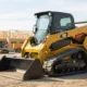 A compact track loader at work on a construction site, with its bucket resting on the ground and other heavy equipment visible in the background.
