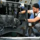 A heavy truck mechanic inspecting and repairing electrical connections on a semi-truck, working carefully with coiled cables and engine components.