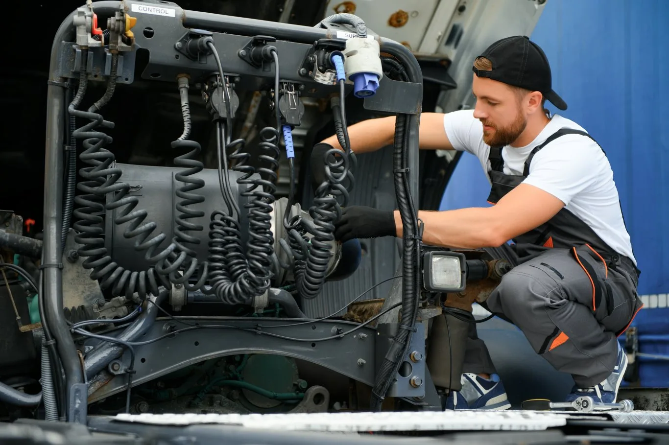A heavy truck mechanic inspecting and repairing electrical connections on a semi-truck, working carefully with coiled cables and engine components.