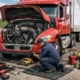 A mechanic performing semi truck repair on a red truck that broke down on the highway, using professional tools and safety cones for roadside service.