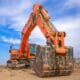 Heavy-duty rental mini excavator on a construction site, positioned for digging and earthmoving work with a large bucket under a clear blue sky.