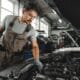 A focused automotive and diesel mechanic at Williams Diesel LLC in Seattle wearing coveralls and protective gloves inspects the complex engine components of a vehicle with the hood raised.