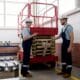A male technician at Williams Diesel LLC wearing a hard hat and coveralls provides hands-on instructions for the safe operation of a red scissors lift rental to a colleague using a tablet in a bright workshop.