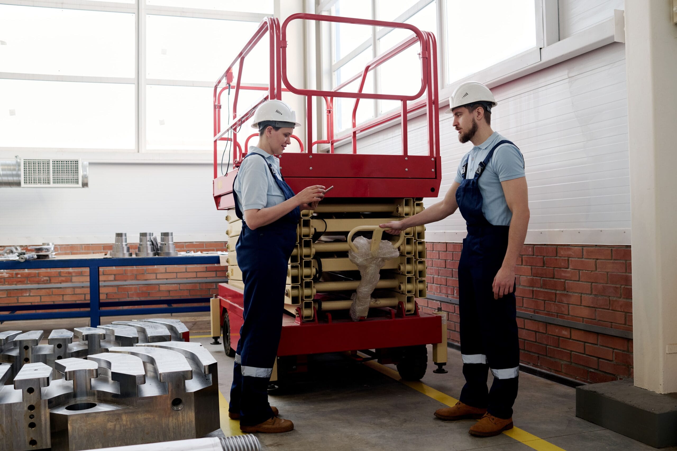 A male technician at Williams Diesel LLC wearing a hard hat and coveralls provides hands-on instructions for the safe operation of a red scissors lift rental to a colleague using a tablet in a bright workshop.