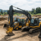 A line of professional excavating equipment, including a tracked excavator and backhoe loaders, parked on a construction site development plot.